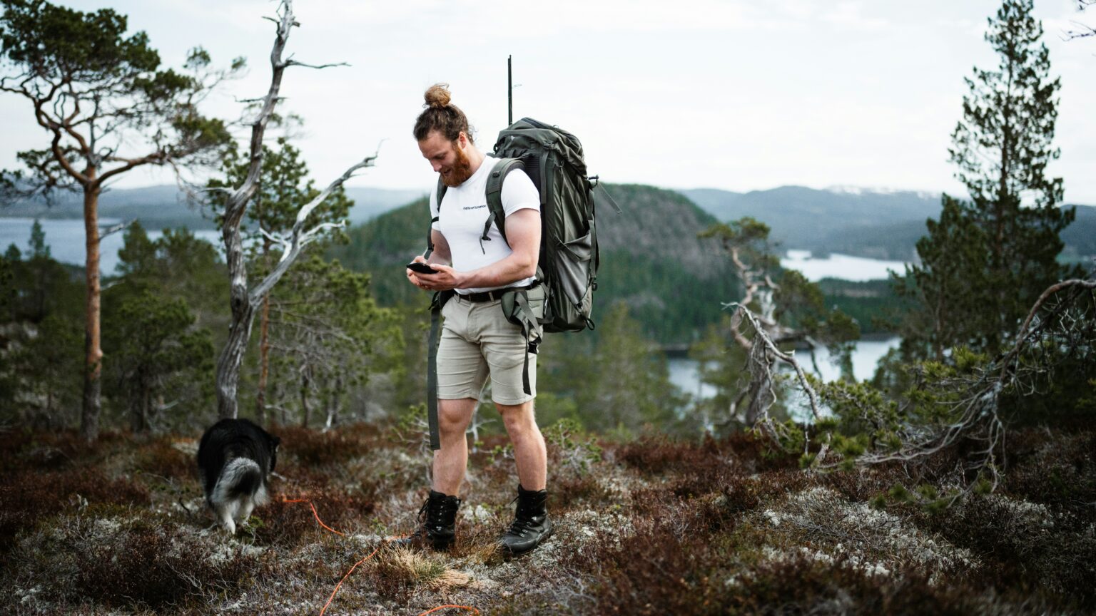 Hiker checking a GPS device on a mountain trail with a backpack and a dog nearby.