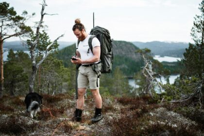 Hiker checking a GPS device on a mountain trail with a backpack and a dog nearby.