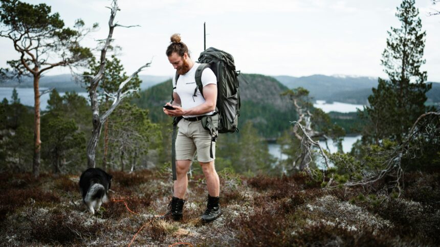 Hiker checking a GPS device on a mountain trail with a backpack and a dog nearby.