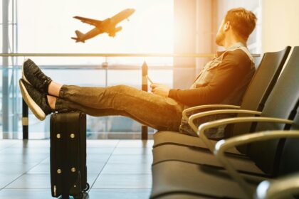 Man waiting at airport gate with feet on suitcase, watching a plane take off before an international flight
