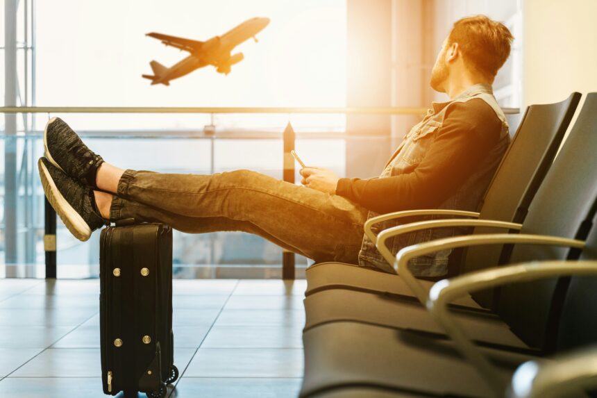 Man waiting at airport gate with feet on suitcase, watching a plane take off before an international flight
