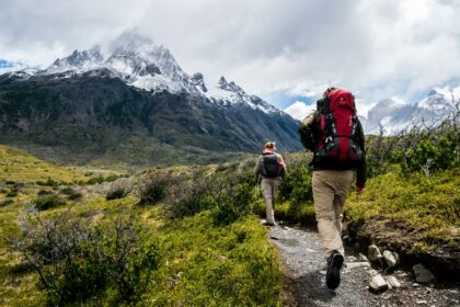 Two hikers walking along a mountain trail with large backpacks and snowy peaks in the background.
